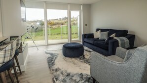 A modern living room with a dark blue sofa, a grey armchair, a patterned rug, and a dining area with a glass table and chairs. - Portnablagh Cottage (Portnablagh)