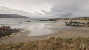 A coastal landscape with a sandy beach, rocky outcrops, and a distant view of houses. - Portnablagh Cottage (Portnablagh)