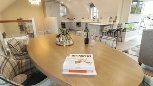 A modern kitchen with white cabinets, a dining area with plaid chairs, and a table with a magazine and wine glasses. - Portnablagh Cottage (Portnablagh)