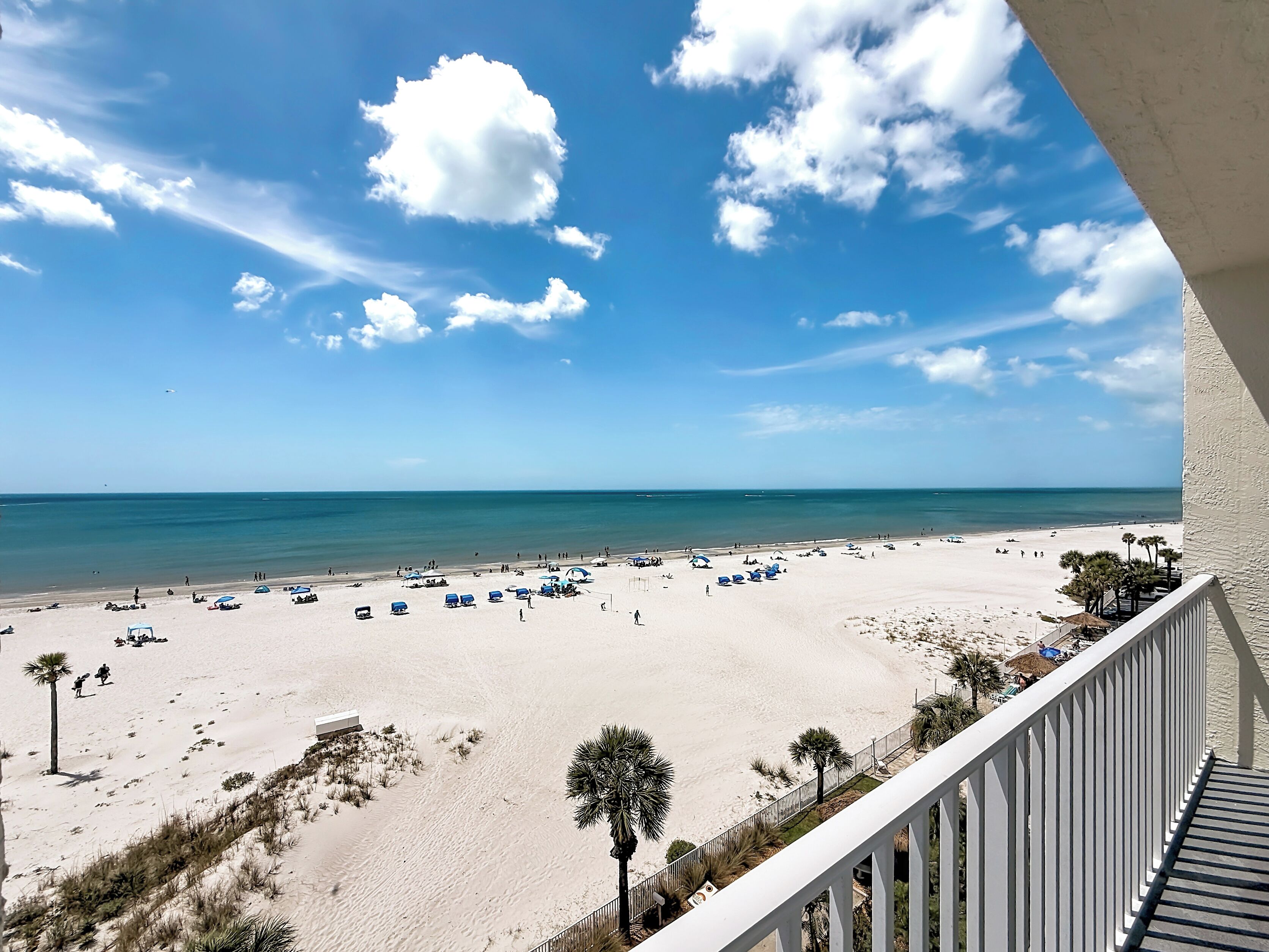 A beach view from a balcony with a railing, palm trees, and a clear sky.