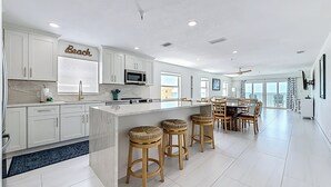 Una cocina moderna con armarios blancos, una isla central con taburetes, una zona de comedor con mesa y sillas, y un ventanal con vistas.