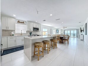 A modern kitchen with white cabinets, a central island with stools, a dining area with a table and chairs, and a large window offering a view. - Amazing Sunset Views! Beachfront unit - Pool - Spa (Madeira Beach)