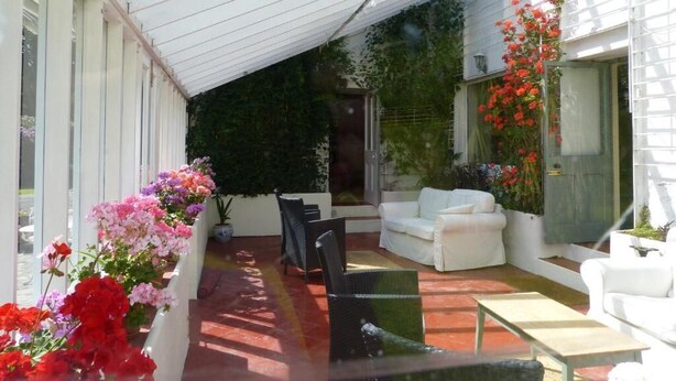 Uma área de estar coberta ao ar livre com móveis brancos, parede de vidro e pérgola branca.