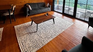 Ein geräumiges Zimmer mit Holzbalkendecke, einer Couch, einem Couchtisch und einem großen Fenster mit Blick ins Freie.