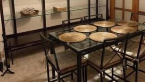 Een eethoek met een tafel met glazen blad, zwarte stoelen en een geweven lamp.