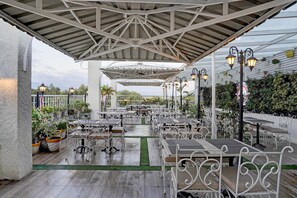 Un patio exterior con mesas y sillas, una zona de estar cubierta e iluminación decorativa.