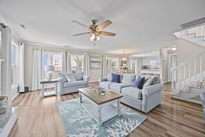 Ein geräumiges Wohnzimmer mit Deckenventilator, zwei grauen Sofas, einem Holztisch und Blick zur Küche im Hintergrund.