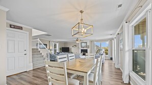 Een ruime woonkamer met een houten eettafel, witte stoelen, een kroonluchter en een trap naar een hoger niveau.