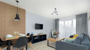 A modern living room with a sofa, dining table, and chairs, a flat-screen TV, and a large window with curtains. - Stylish Apartment | Balcony, Parking, Wellness Area (Dziwnów)