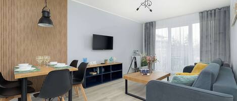 A modern living room with a sofa, dining table, and chairs, a flat-screen TV, and a large window with curtains.