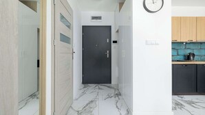 A hallway with a dark door, a clock on the wall, and a kitchen area with wooden cabinets and a blue backsplash. - Stylish Apartment | Balcony, Parking, Wellness Area (Dziwnów)