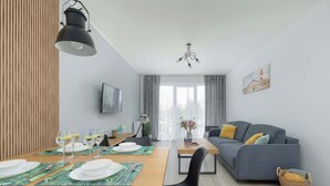 A modern living room with a dining area, grey sofa, and a TV mounted on the wall. - Stylish Apartment | Balcony, Parking, Wellness Area (Dziwnów)