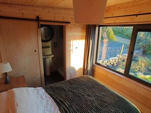 Un dormitorio de cabaña de madera con cama, mesita de noche, lámpara, ventana y vistas al exterior.