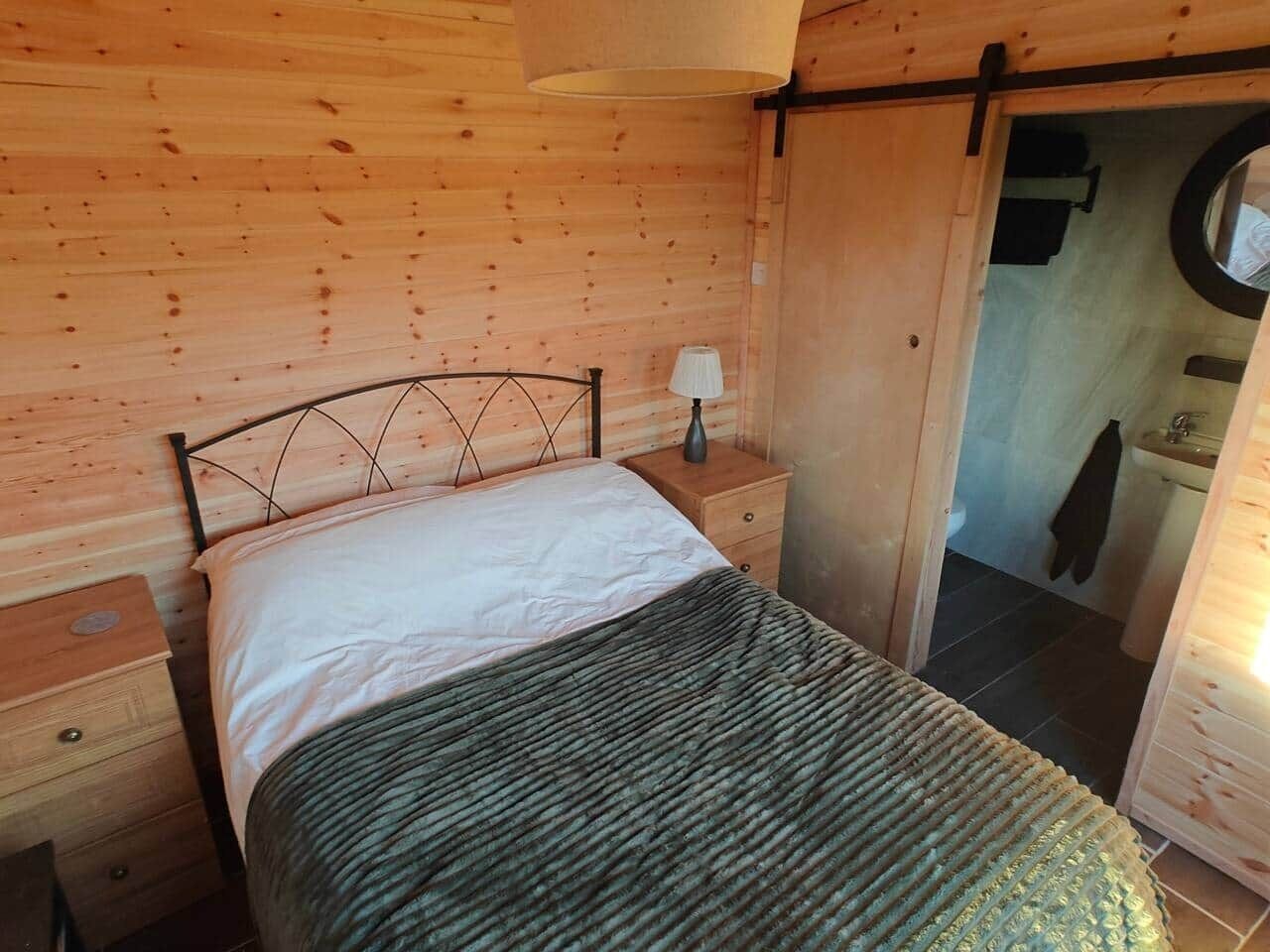 Quarto de cabana de madeira com uma cama, uma mesa de cabeceira, um abajur e uma porta que dá para o banheiro.
