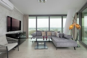 Ein modernes Wohnzimmer mit einem großen Flachbildfernseher, einem grauen Ecksofa, einem Glastisch und einem großen Fenster mit Blick auf die Stadt.