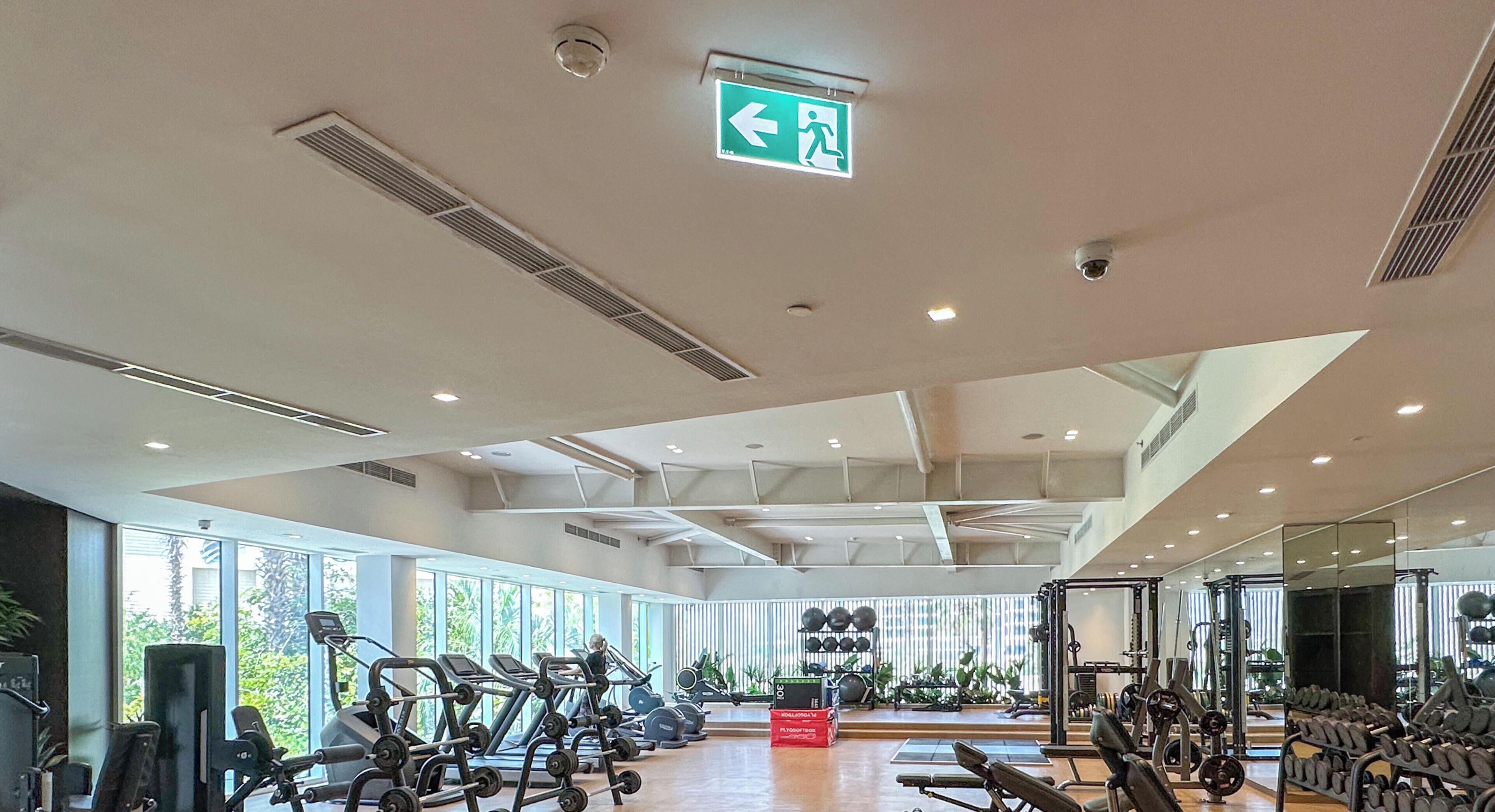 A spacious gym with various exercise equipment, large windows, and an emergency exit sign.