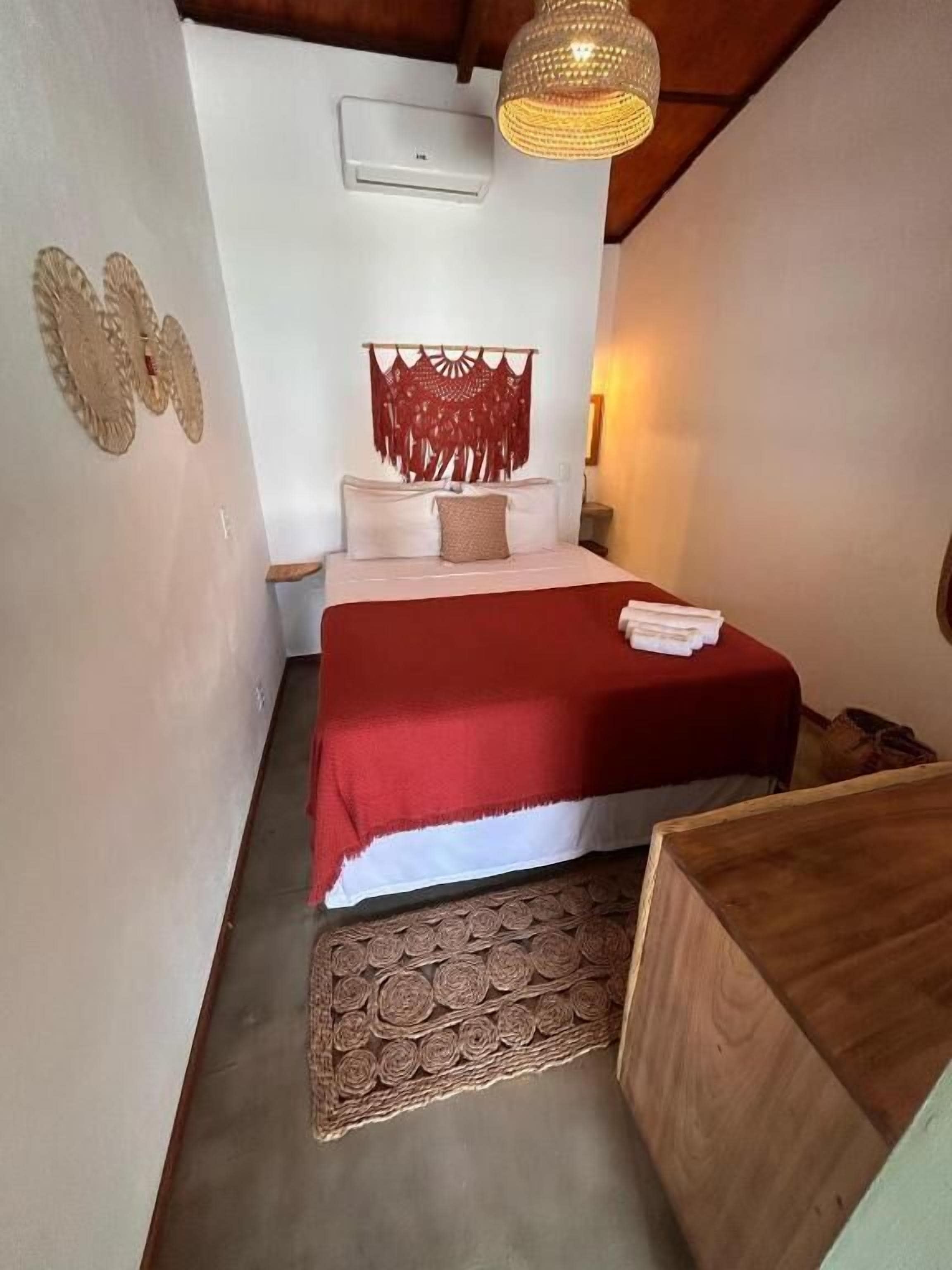 Un dormitorio con una cama, una colcha roja, un cabecero de madera, una lámpara colgante y una alfombra estampada.