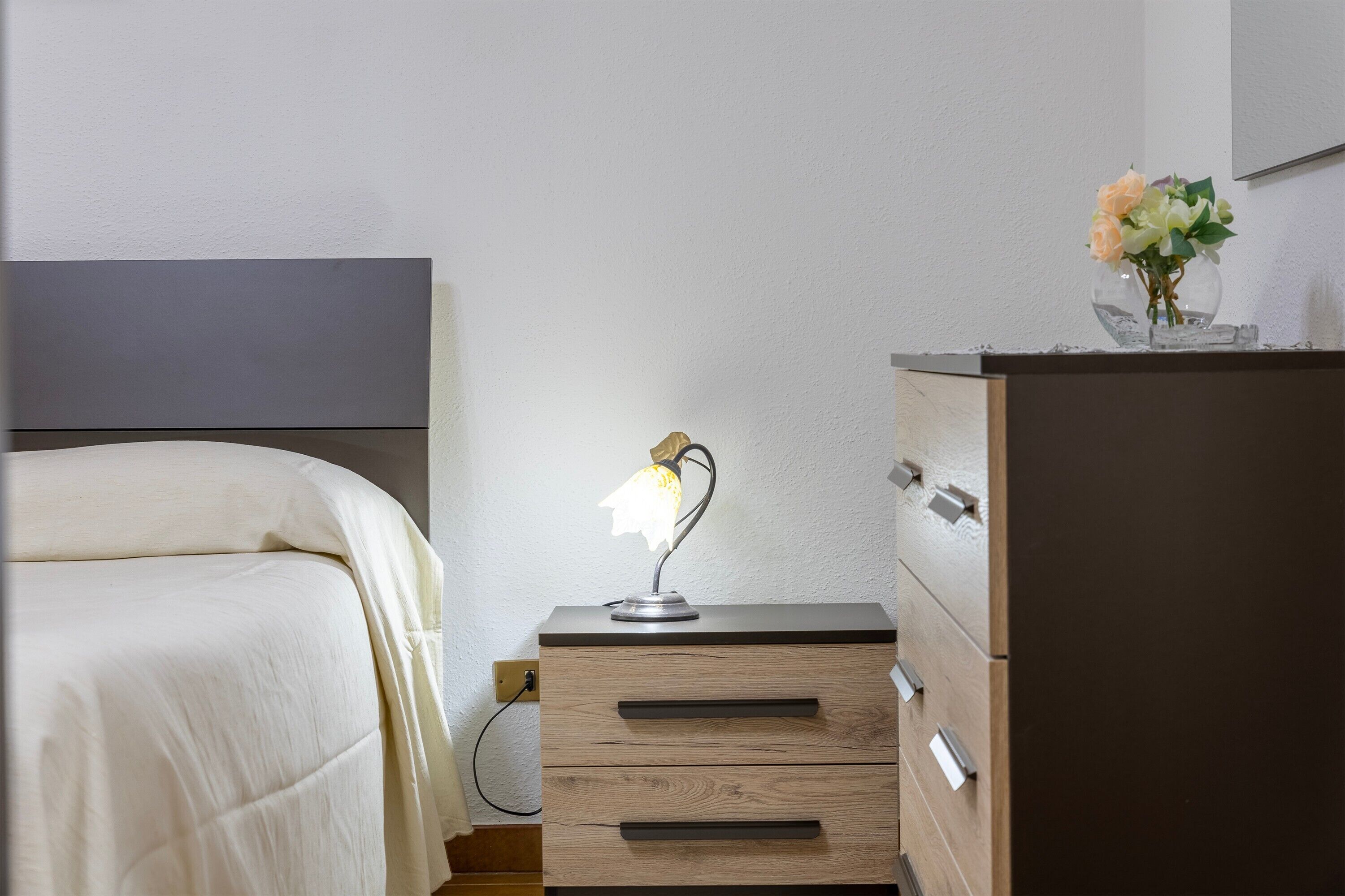Un dormitorio con una cama, una lámpara de noche, un tocador y un jarrón con flores sobre él.
