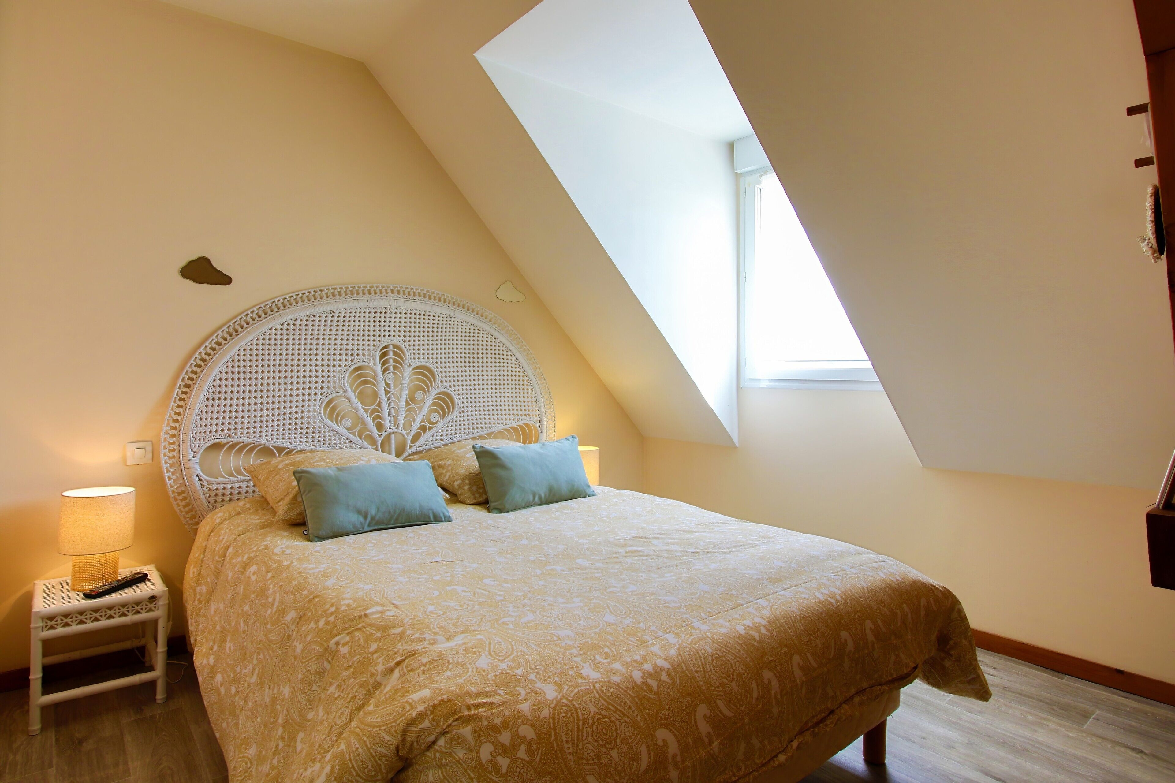 Een slaapkamer met een schuine kap, een bed met beddengoed met een patroon, twee kussens, een nachtkastje met een lamp en een raam dat natuurlijk licht binnenlaat.