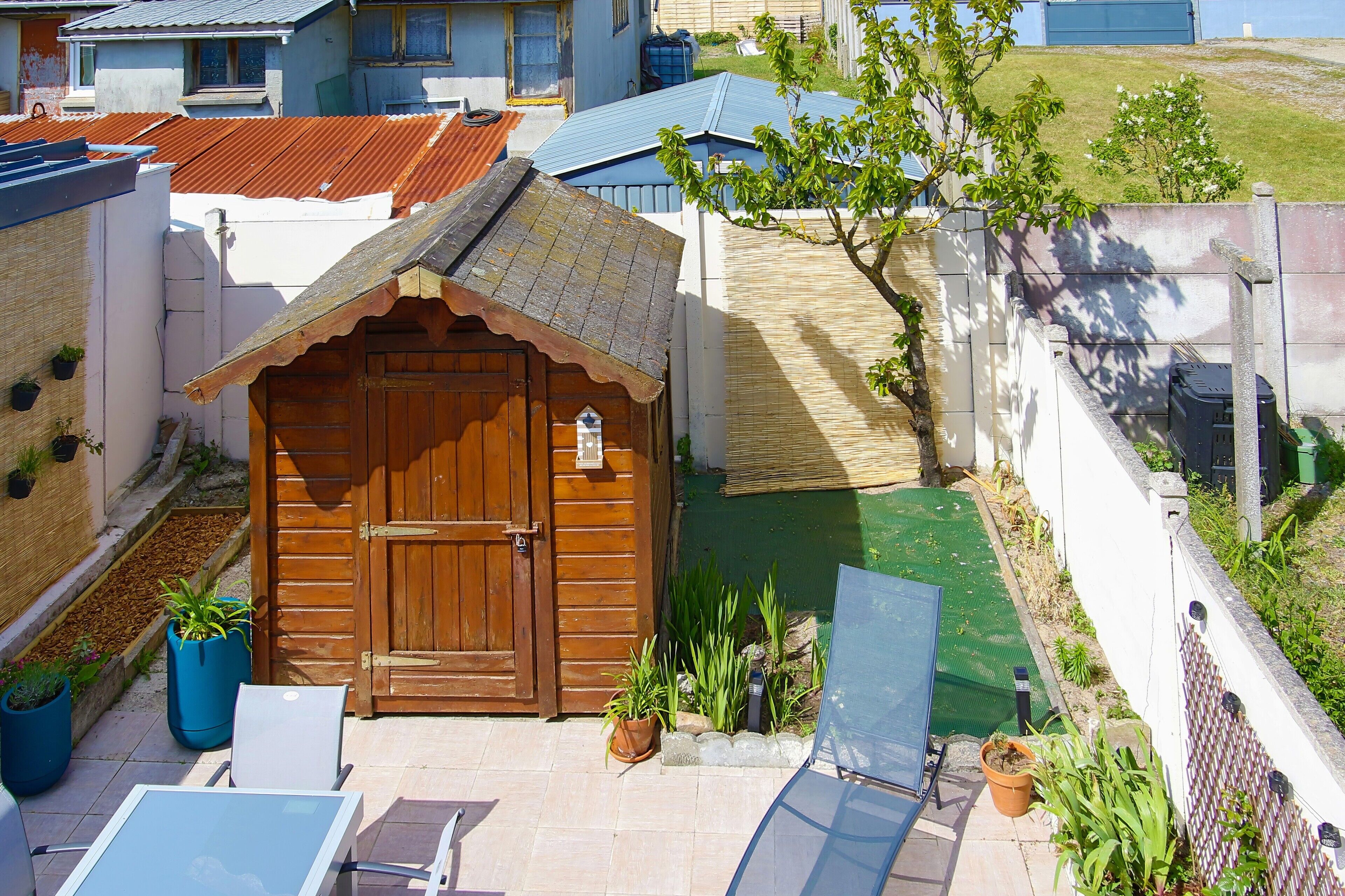A wooden shed with a door, surrounded by potted plants, a small garden, and a patio with a table and chairs.