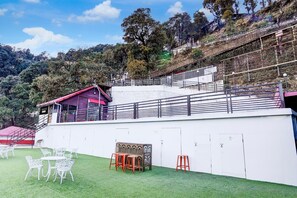 Une terrasse avec des chaises et des tables en métal blanc, un bâtiment au toit rouge et un mur blanc avec des portes.