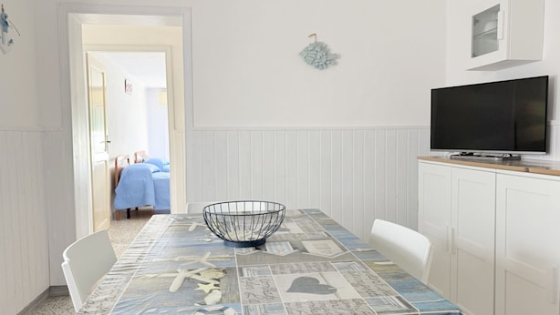 Een eethoek met een tafel bedekt met een tafelkleed met dessin, een metalen kom, witte stoelen, een televisie op een kast en een deur naar een andere kamer.
