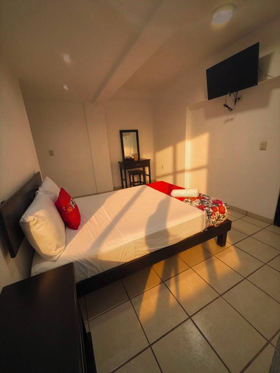 A single bed with a red pillow, a bedside table, a small desk with a chair, and a mounted TV on the wall.