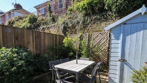 Een houten tafel met stoelen in een tuin, met een houten hek en een tuinhuis op de achtergrond.