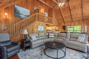 A spacious living area with a large L-shaped sectional sofa, a round coffee table, a staircase with a railing, and a ceiling fan. - Lake Days & Sunset Nights - Beautiful 5 bedroom house on Norris Lake w/ dock! (Speedwell)
