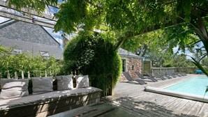 A wooden deck with a pool, a stone building, and a seating area with cushions.