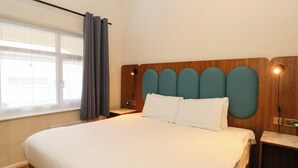 A hotel room with a large bed, a wooden headboard, a nightstand, a window with curtains, and a wall-mounted light. - Stag Lodge (Chorley)