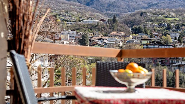 A balcony with a table, a bowl of fruit, and a view of a town with buildings and mountains in the background. - Bellavista Saint-Vincent (Saint-Vincent)