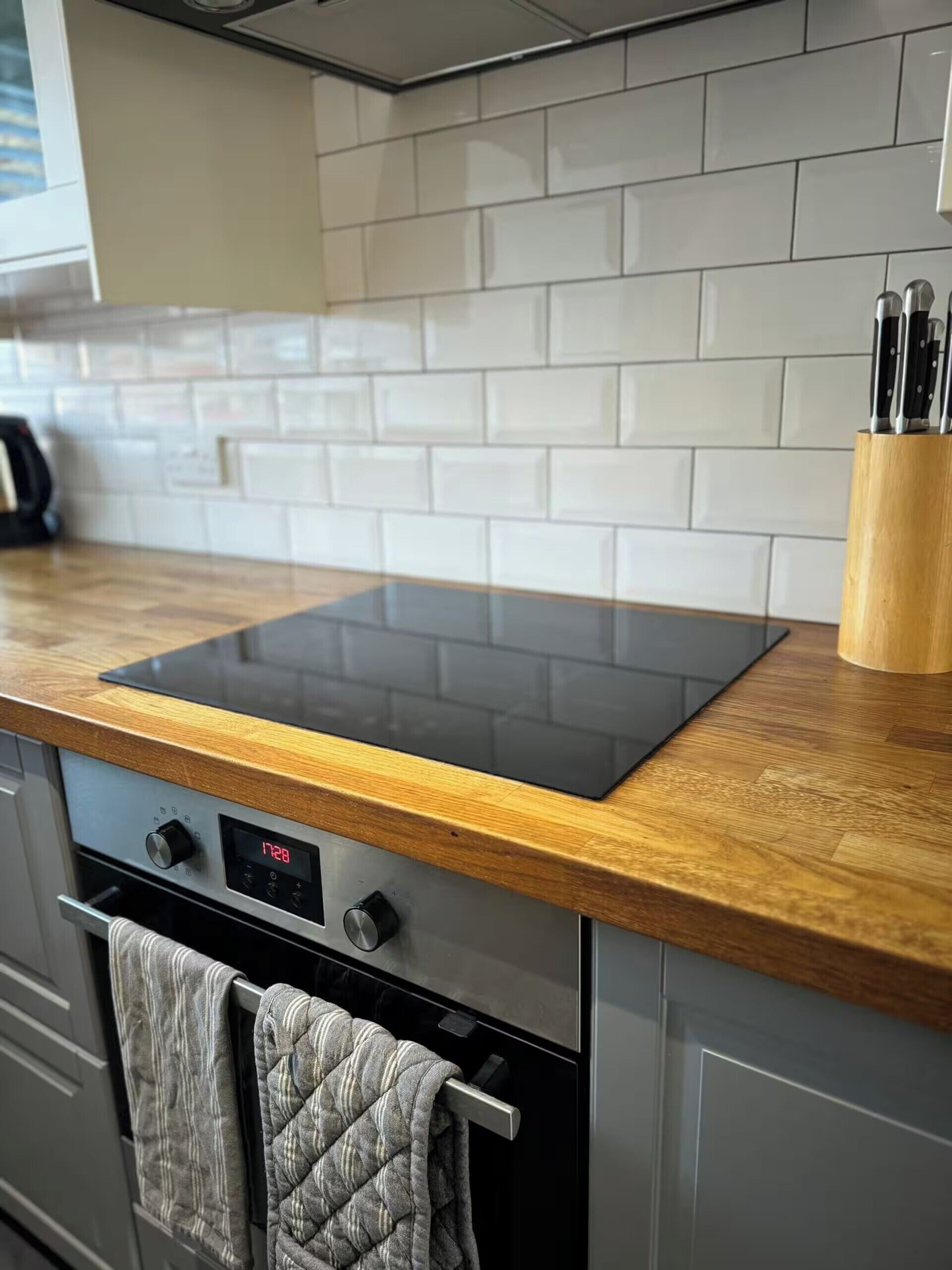 A kitchen with a black induction cooktop, wooden countertop, stainless steel oven, and a tiled backsplash.