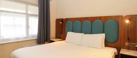 Une chambre d’hôtel avec un grand lit, une tête de lit en bois, une table de chevet, une fenêtre avec des rideaux et un lampadaire fixé au mur.