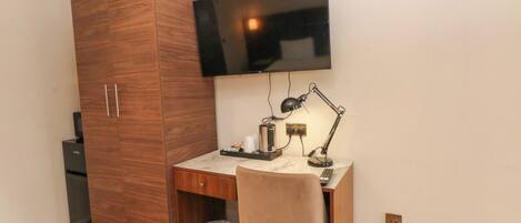 Un dormitorio con un armario de madera, un escritorio con silla, un televisor montado en la pared y un cesto de basura.