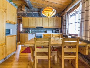 A wooden interior with a dining table and chairs, a hanging light fixture, and a window with curtains.