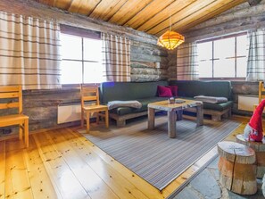 A rustic cabin interior with a wooden floor, a green sofa, a wooden chair, a small table, and a hanging light fixture.