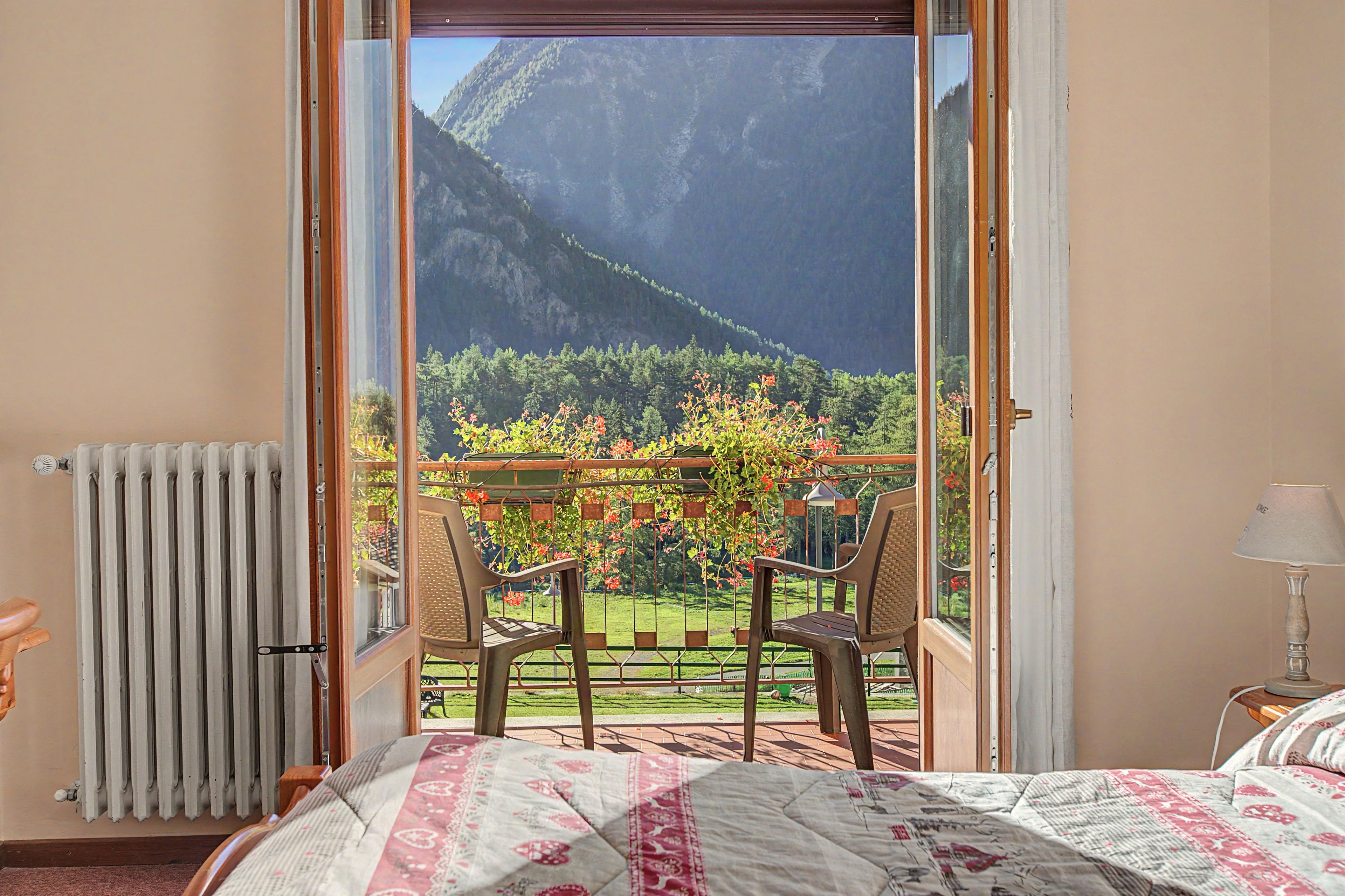 A room with a bed, a radiator, a table with chairs, and a view of a mountainous landscape through an open door.