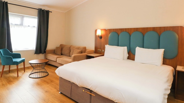 Quarto de hotel com uma cama grande, um sofá, uma cadeira, uma mesinha e uma janela com cortinas.