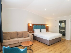 A hotel room with a bed, a sofa, a small round table, and a bathroom with a sink. - Hedgehog Lodge (Chorley)