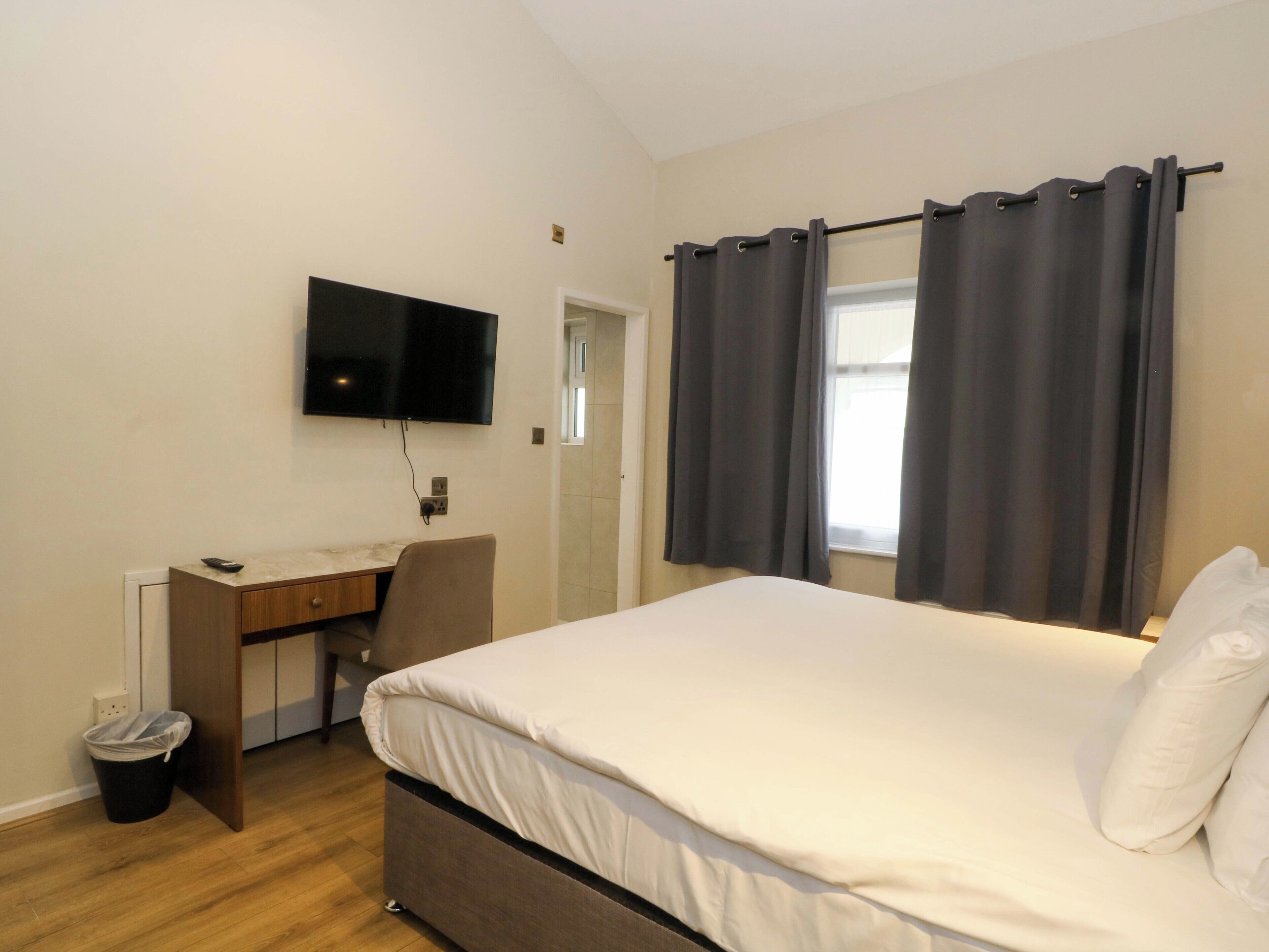 A bedroom with a bed, a desk, a chair, a TV, and a window with curtains.
