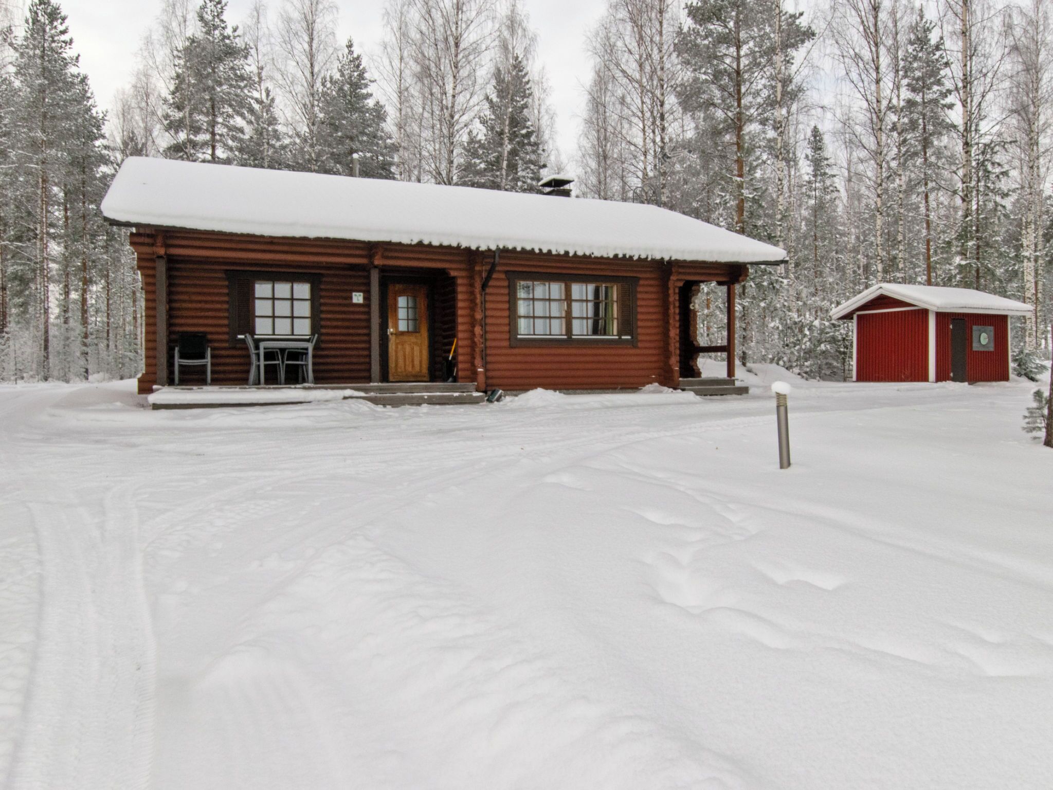 Une cabane en bois avec un porche, une remise rouge séparée et un paysage de forêt enneigée en arrière-plan.