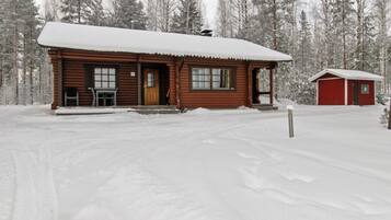Une cabane en bois avec un porche, une remise rouge séparée et un paysage de forêt enneigée en arrière-plan.
