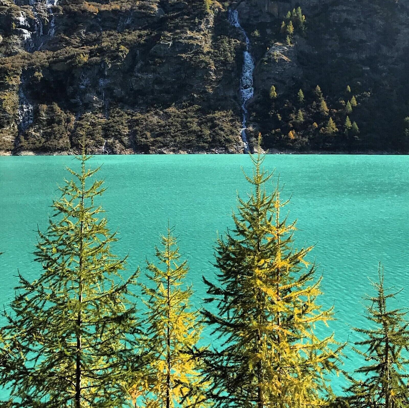A mountainous landscape with a clear turquoise lake, trees in the foreground, and a waterfall in the background.