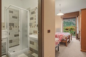 A bathroom with a shower, toilet, and sink. There is a bed with a view of a garden through a window. - "Room 9" with Mountain View, Shared Terrace in Bionaz (Dzovennoz)