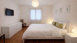 Ein Schlafzimmer mit einem Bett, Nachttischen, einem Sessel, einem Fernseher und einem Fenster mit Vorhängen.