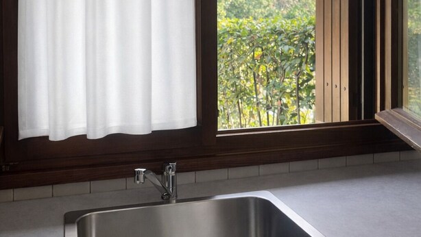 Eine Küche mit Spüle aus Edelstahl, einem Fenster mit weißem Vorhang und Blick auf Bäume und Grünflächen.