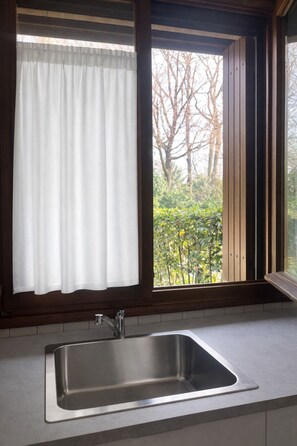 A kitchen with a stainless steel sink, a window with a white curtain, and a view of trees and greenery outside. - Gaidon House, an oasis of peace in the green within walking distance of downtown Bassano (Bassano del Grappa)