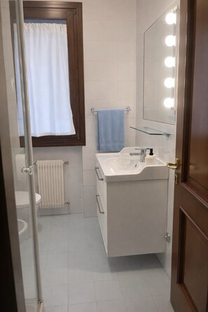 Un bagno con lavandino bianco, specchio illuminato, porta asciugamani e finestra con tenda.