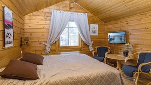 Quarto de cabana de madeira com uma cama, duas cadeiras, uma mesa pequena, uma TV e uma janela com cortinas.